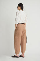 A woman is shown from the back wearing a white blouse, tan wide-leg cropped trousers, a thin brown belt, and black mule sandals against a plain light gray studio background.