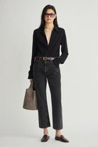 A woman modeling a black long-sleeve button-up shirt, black cropped jeans, a brown belt, and dark brown loafers, holding a taupe-gray tote bag against a light gray studio background.