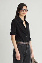 A woman modeling a black long-sleeve button-up shirt with rolled sleeves, paired with dark gray jeans, a brown belt, and black sunglasses, against a light gray studio background.