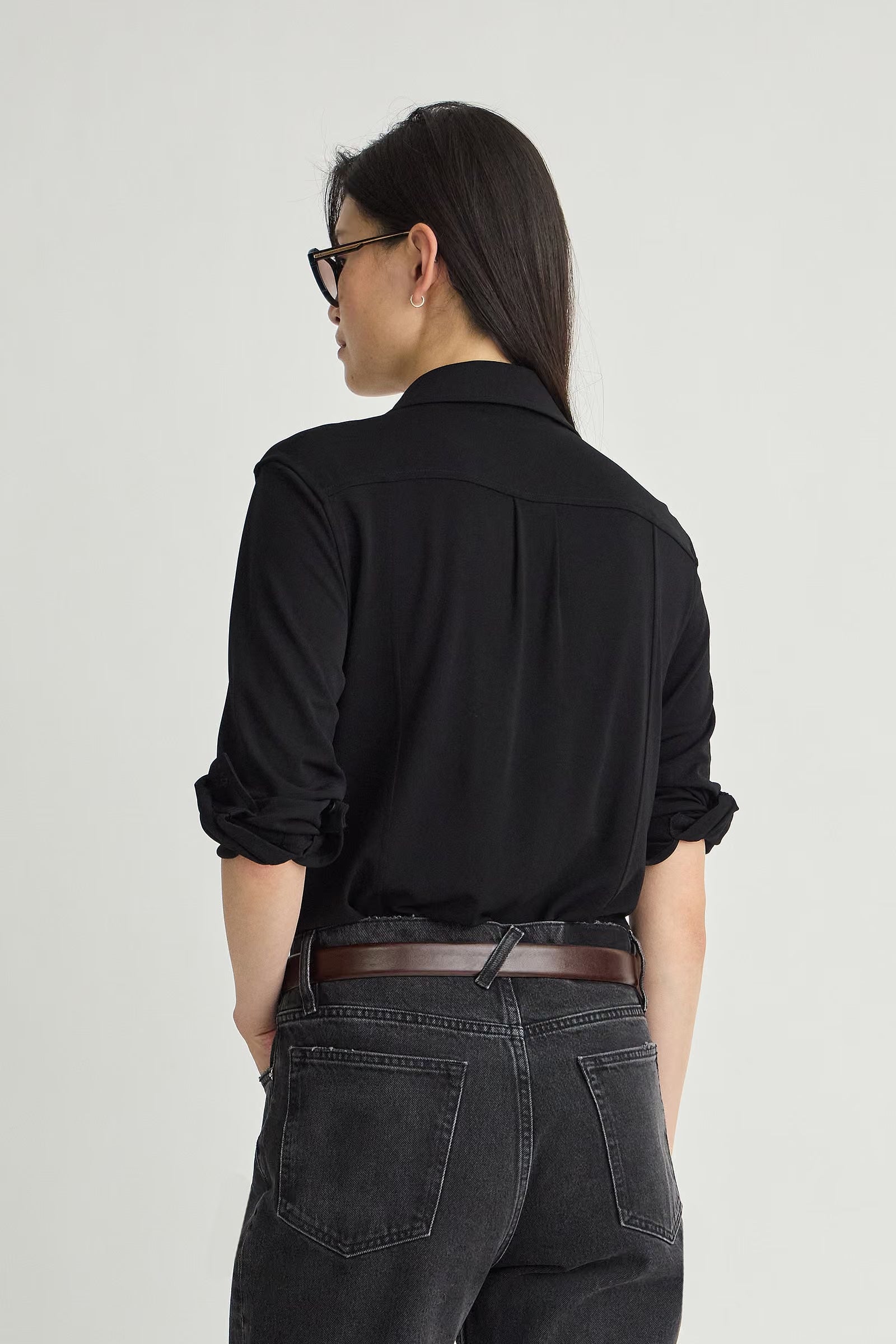 A woman shown from the back wearing a black long-sleeve button-up shirt, dark gray jeans, and a brown belt, photographed against a light gray studio background.