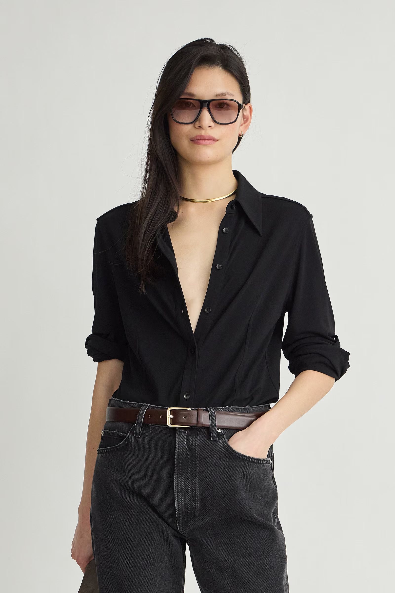 A woman modeling a black long-sleeve button-down shirt with rolled sleeves, paired with dark gray jeans, a brown belt, and black sunglasses, against a light gray studio background.