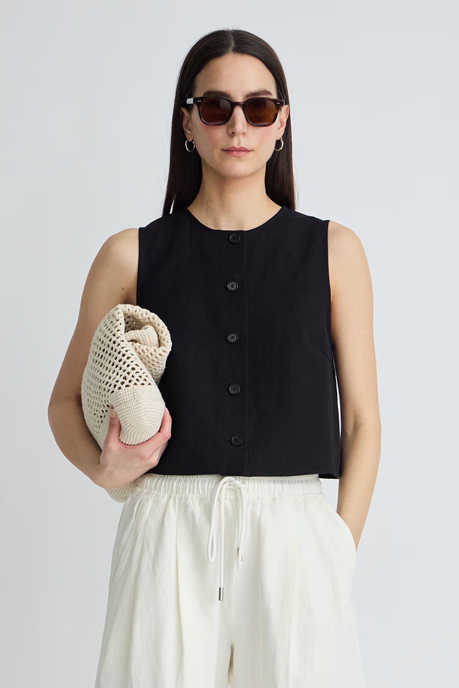 Woman wearing a black sleeveless top and white pants, holding a beige knitted bag against a plain background.