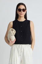 Woman wearing a black sleeveless top and white pants, holding a beige knitted bag against a plain background.