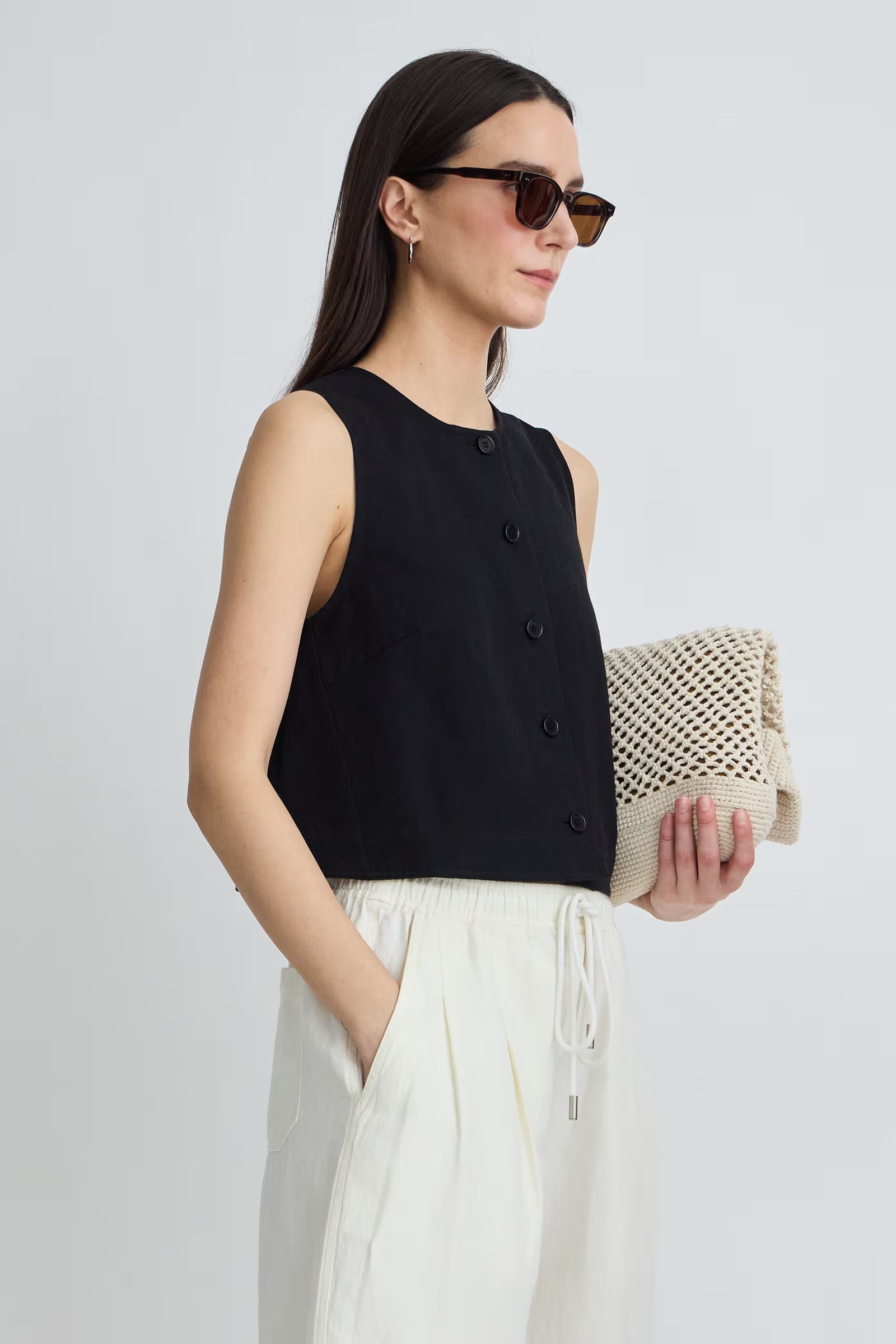 Woman wearing a black sleeveless top and white pants, holding a beige bag against a light gray background