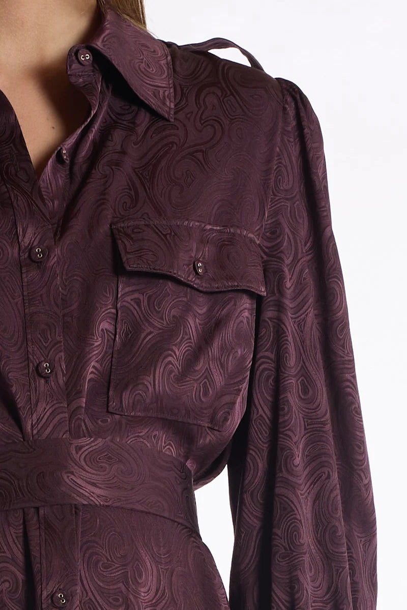 A close-up of a dark plum purple long-sleeve shirt dress with a swirling jacquard pattern, featuring a button-down front, chest pocket, and matching waist tie.