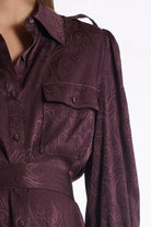 A close-up of a dark plum purple long-sleeve shirt dress with a swirling jacquard pattern, featuring a button-down front, chest pocket, and matching waist tie.
