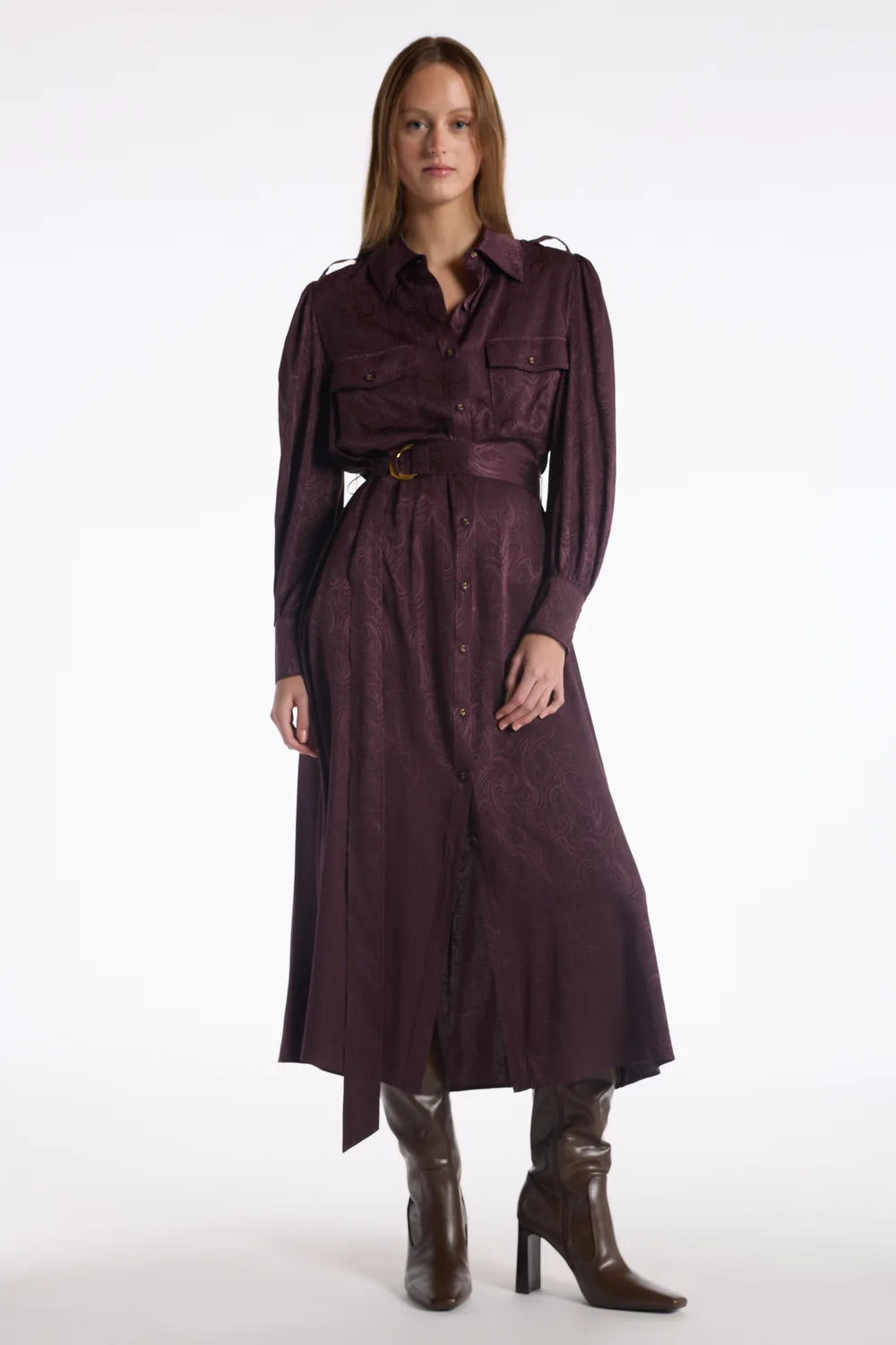 A woman modeling a long, dark plum-burgundy midi shirt dress with a matching waist belt and brown heeled ankle boots, photographed against a seamless white studio background.