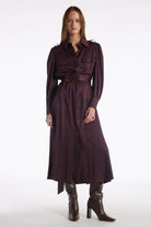 A woman modeling a long, dark plum-burgundy midi shirt dress with a matching waist belt and brown heeled ankle boots, photographed against a seamless white studio background.