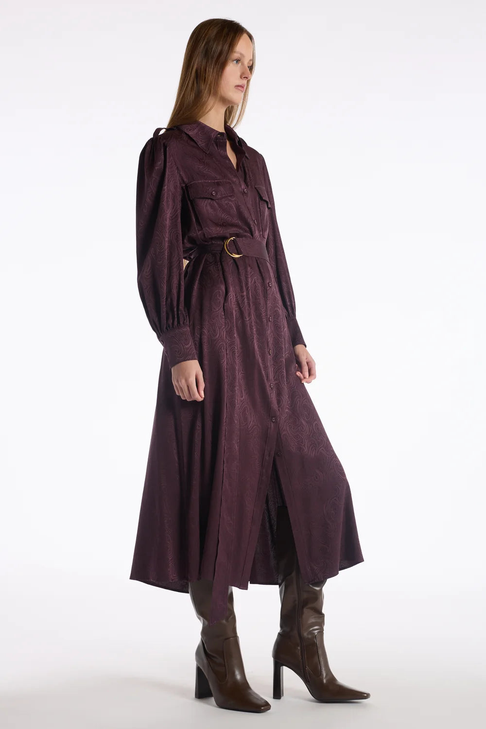 A model wearing a long, dark plum-purple belted coat with puffed sleeves, styled with brown leather heeled boots against a plain white studio background.