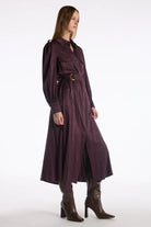 A model wearing a long, dark plum-purple belted coat with puffed sleeves, styled with brown leather heeled boots against a plain white studio background.