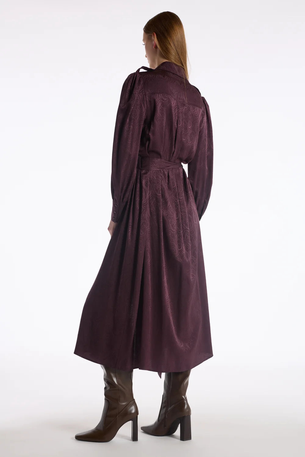 A woman shown from the back wearing a long, dark plum-burgundy belted coat or dress with long sleeves, paired with brown heeled ankle boots, against a plain white studio background.