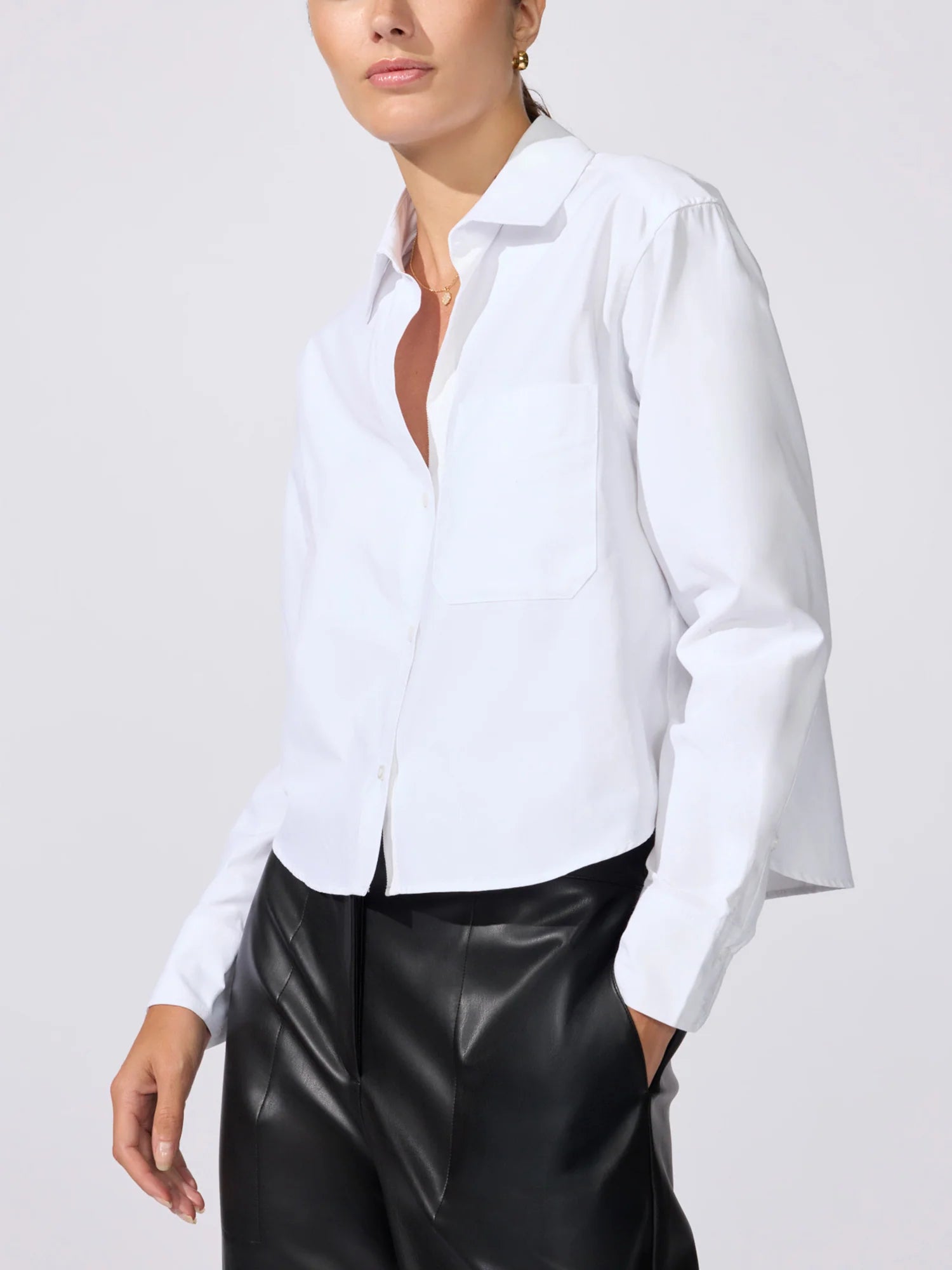 A woman wearing a white long-sleeve button-up shirt with a chest pocket and black leather-look pants, photographed against a light gray studio background.