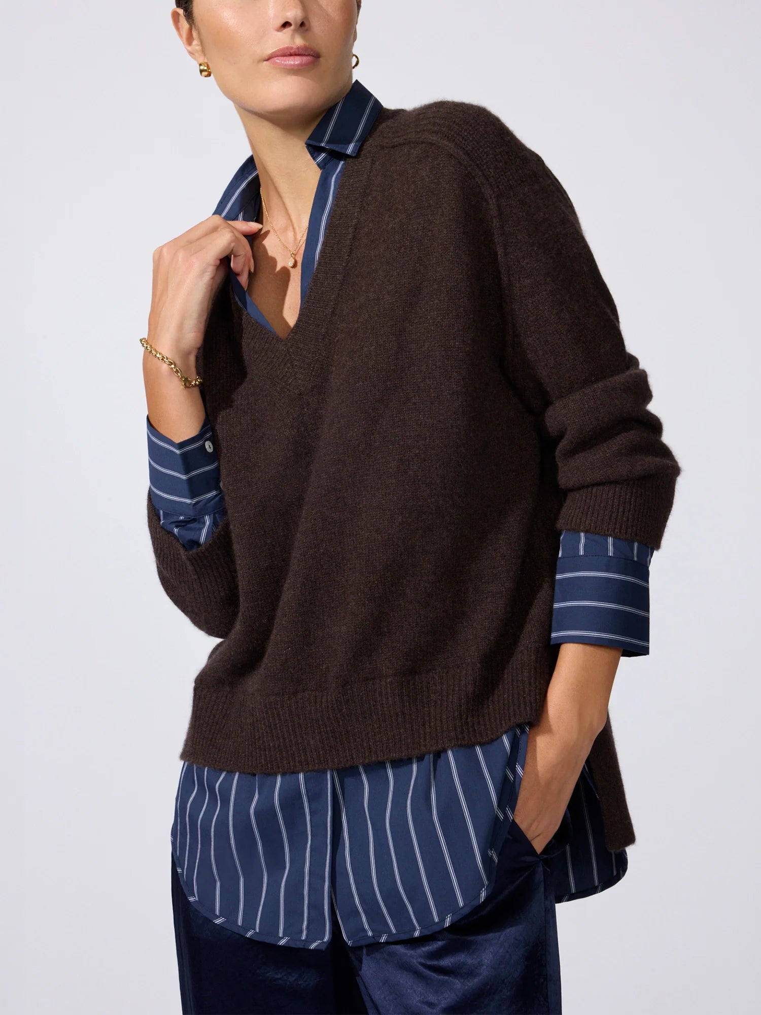 A woman models a dark brown knit sweater layered over a blue and white striped collared shirt, paired with navy trousers, against a light gray studio background.