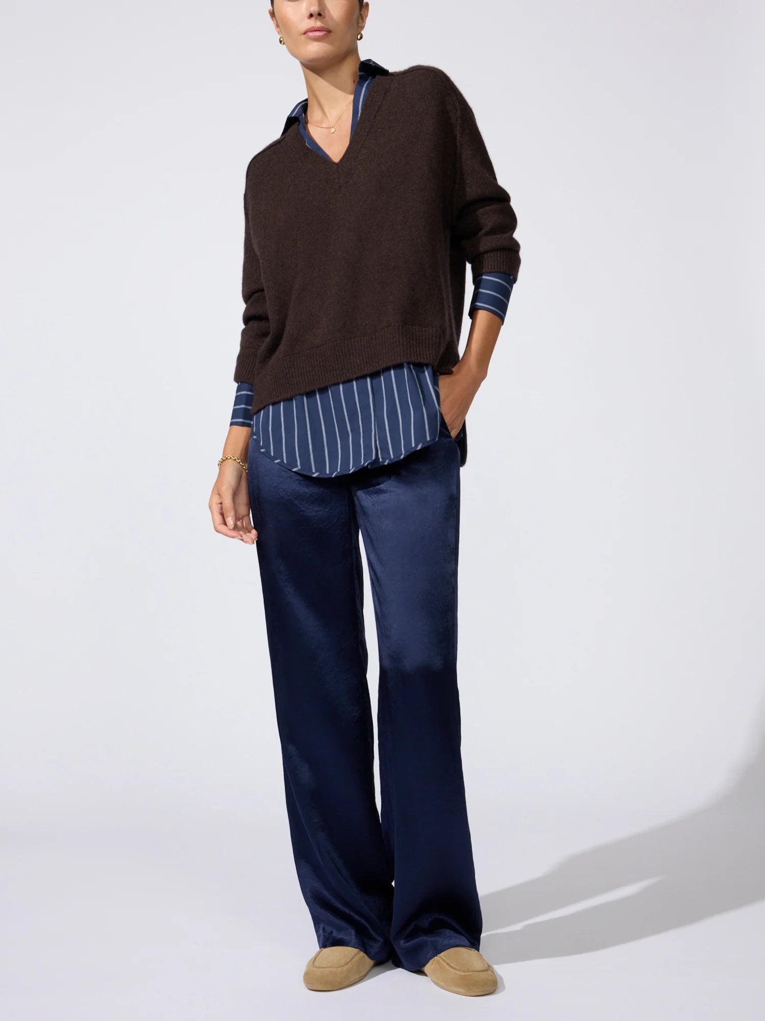 A model wearing a dark brown v-neck knit sweater layered over a blue and white striped shirt, paired with navy blue wide-leg trousers and tan suede shoes, against a plain light gray studio background.