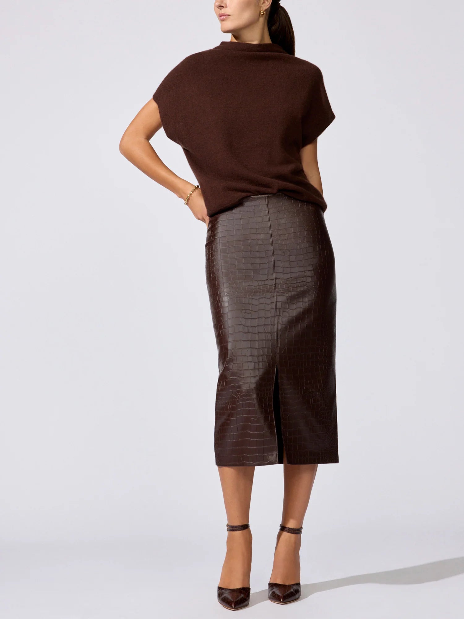 A woman modeling a dark brown short-sleeve knit mock-neck top and a dark brown faux-croc leather midi skirt, paired with brown high heels, against a light gray studio background.