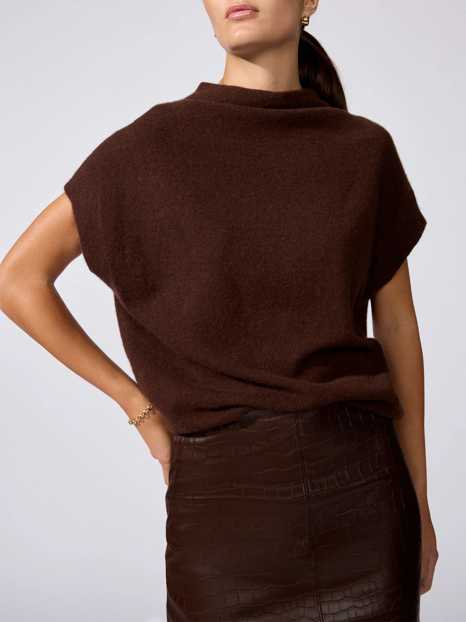A woman wearing a dark brown short-sleeve knit top and a dark brown faux-leather skirt, shown from the back against a light gray studio background.