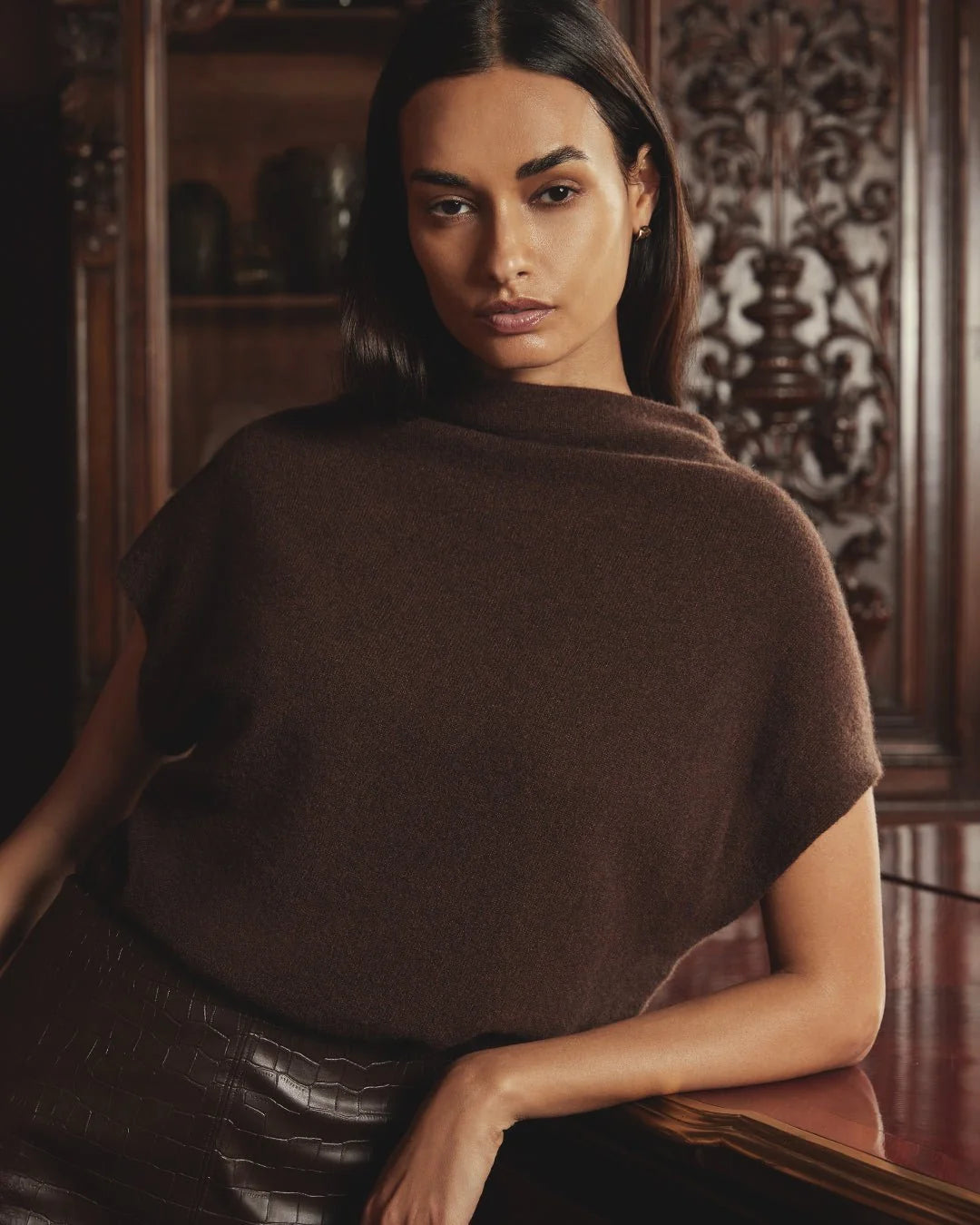 A woman wearing a dark brown knit draped poncho-style top, posed in front of a dark wood cabinet.