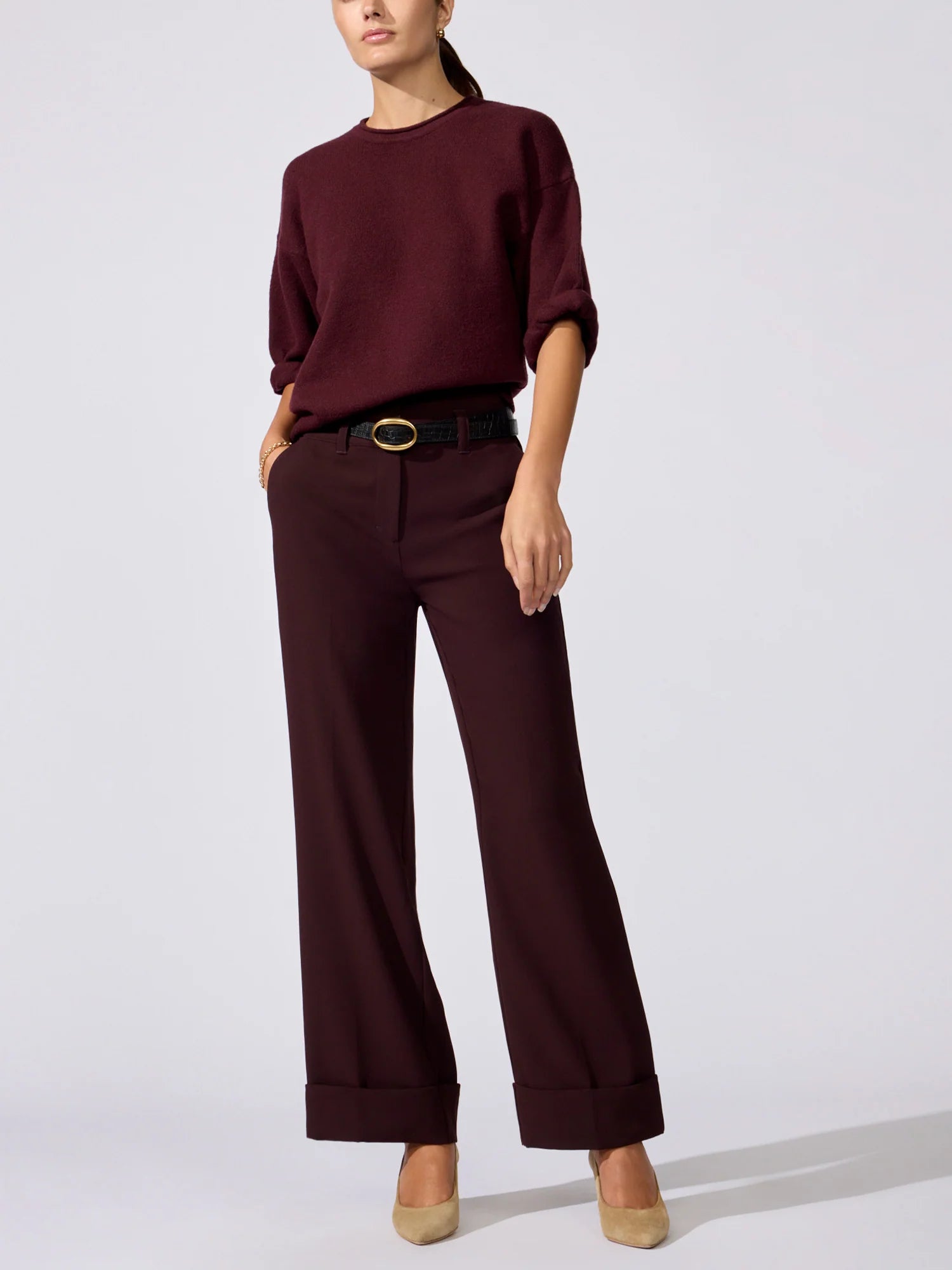 A model wearing a dark burgundy crewneck sweater and matching wide-leg trousers with a black belt, styled with beige heels against a light gray studio background.