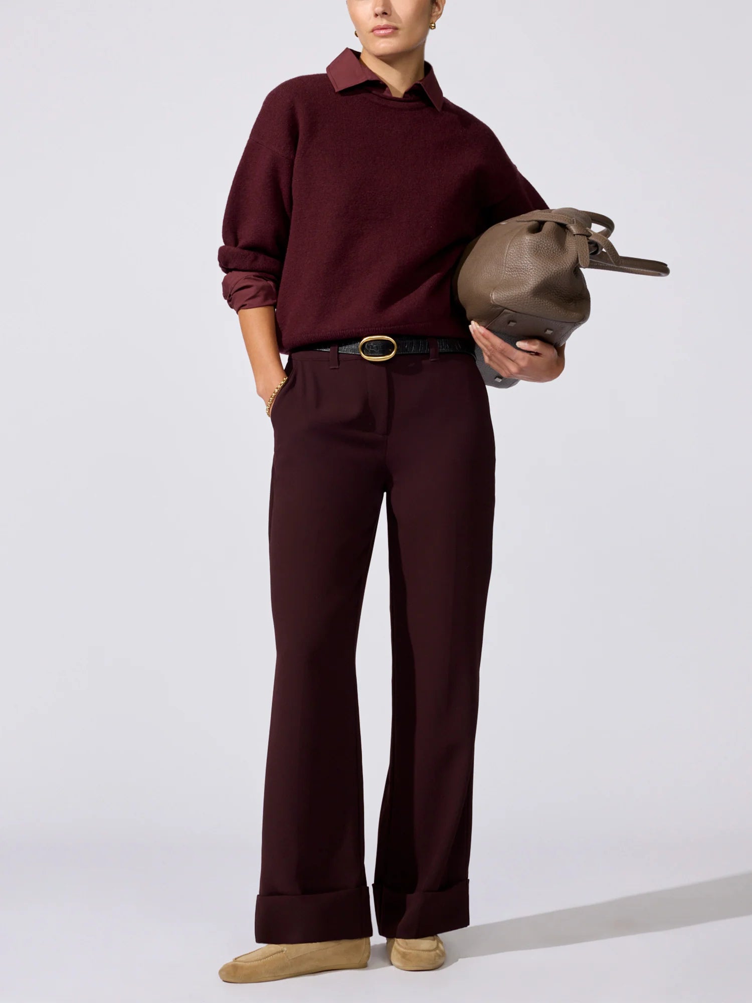 A model wearing a dark burgundy knit polo sweater and matching wide-leg trousers, styled with a black belt, tan shoes, and holding a brown leather bag against a light gray studio background.