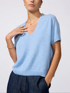 A woman wearing a light blue short-sleeve knit V-neck sweater and dark blue trousers, photographed against a clean light gray studio background.