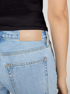 Close-up of the back waistband of light-wash blue jeans with a tan leather patch embossed with 'ARELIE OTH + FRANK', worn with a black top against a light gray background.