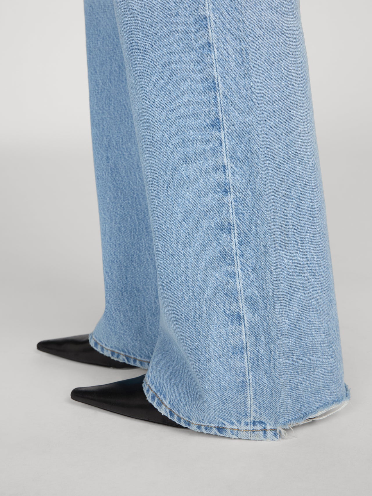 Close-up of wide-leg light blue denim jeans with a raw frayed hem, paired with black pointed-toe flats, shown against a neutral light gray background.