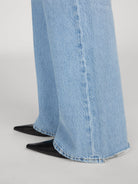 Close-up of wide-leg light blue denim jeans with a raw frayed hem, paired with black pointed-toe flats, shown against a neutral light gray background.