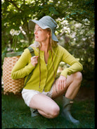 Woman in a green jacket and white shorts squatting outdoors with a basket and green hat.