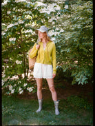 Woman in a garden wearing a yellow cardigan, white skirt, and gray rain boots.