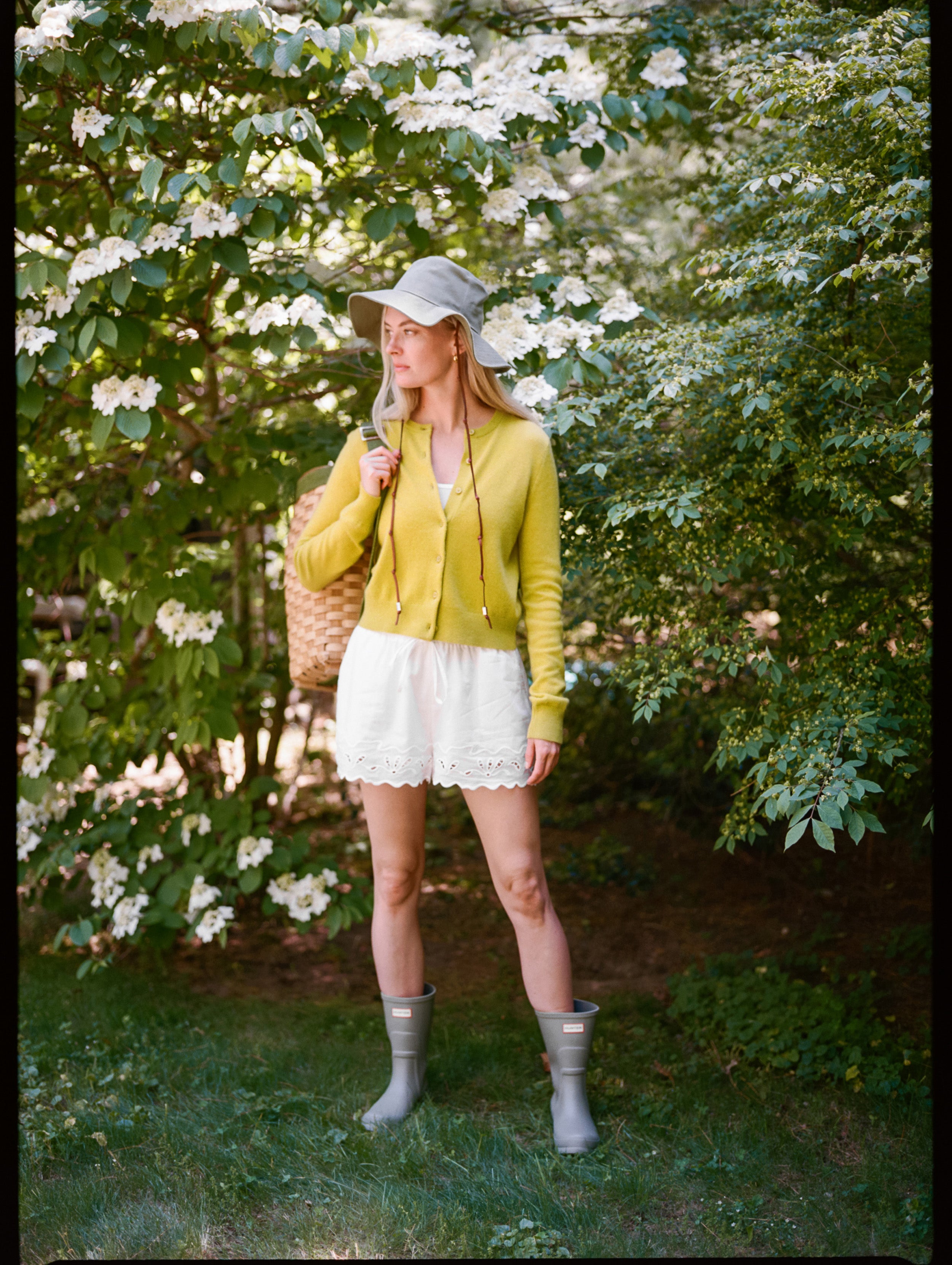 Woman in a garden wearing a yellow cardigan, white skirt, and gray rain boots.