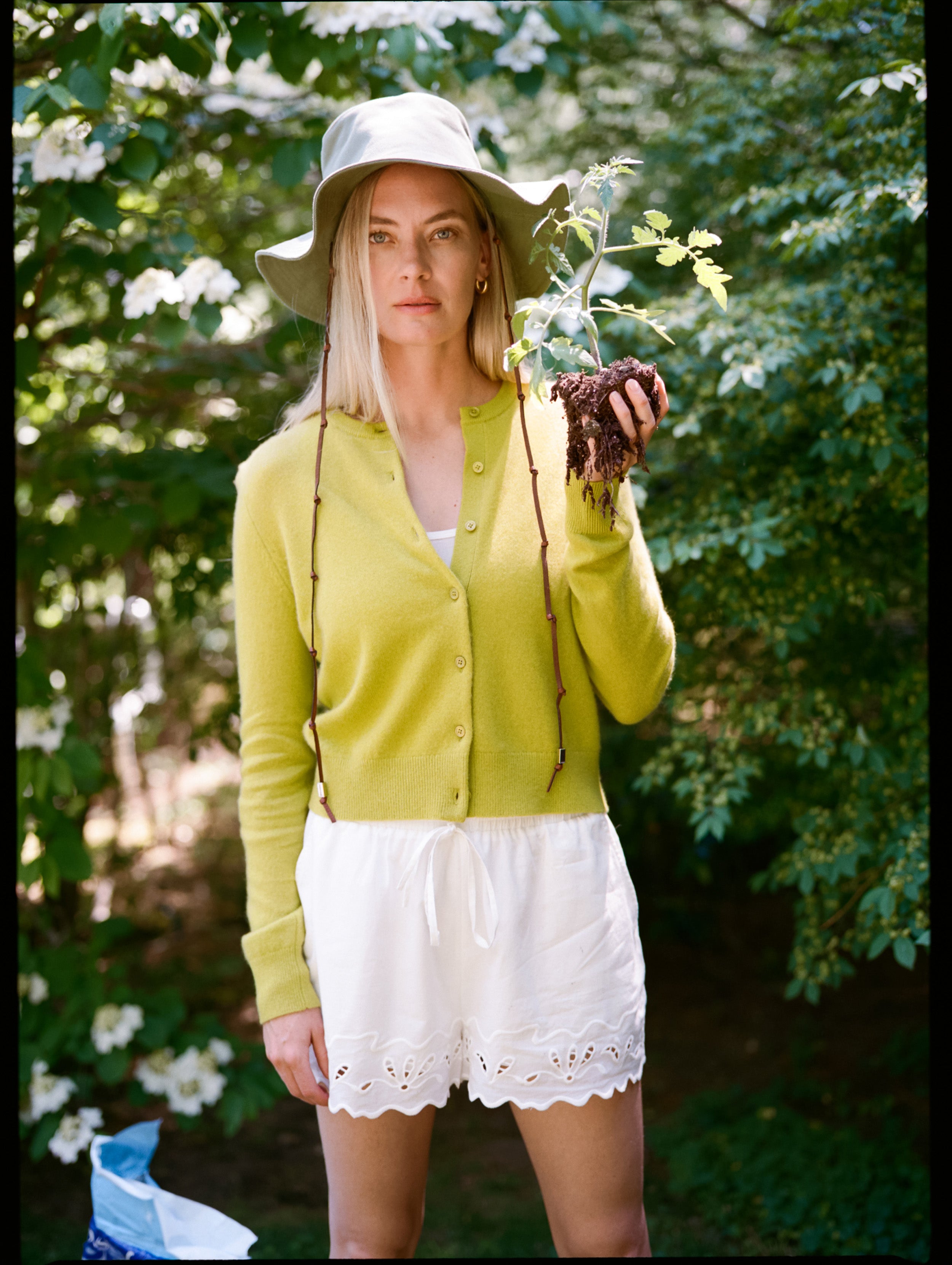Woman in a yellow cardigan and white shorts holding a small plant outdoors.