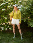 Woman in a garden wearing a green cardigan, white shorts, and green rain boots.