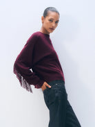 A woman modeling a deep burgundy knit sweater with fringe detailing on the sleeve, paired with dark wide-leg jeans against a clean white studio background.