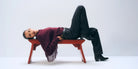 A model lies on a red wooden bench against a light gray studio background, wearing a burgundy sweater with fringe, black pants, and black boots.