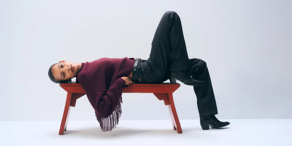 A model lies on a red wooden bench against a light gray studio background, wearing a burgundy sweater with fringe, black pants, and black boots.