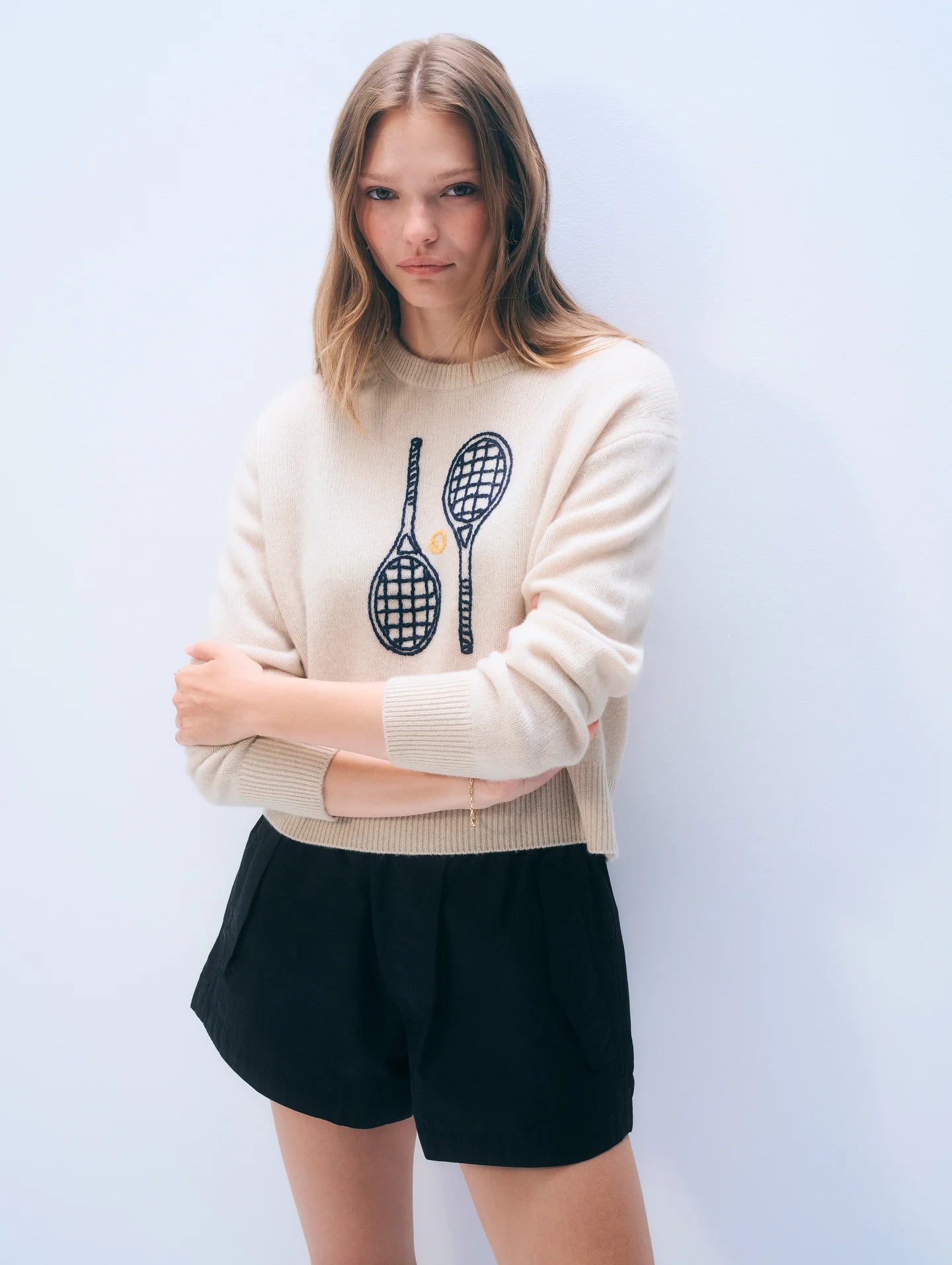 Woman wearing a cream sweater with tennis racket design and black shorts on a light blue background