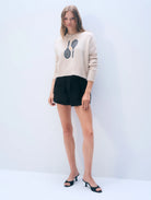 Woman wearing a beige sweater with a graphic design and black shorts on a white background