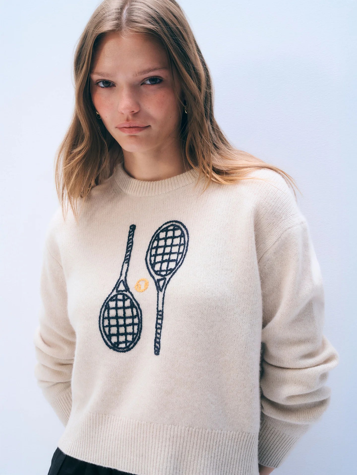 Woman wearing a cream sweater with tennis racket design on a light blue background