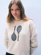 Woman wearing a cream sweater with tennis racket design on a light blue background