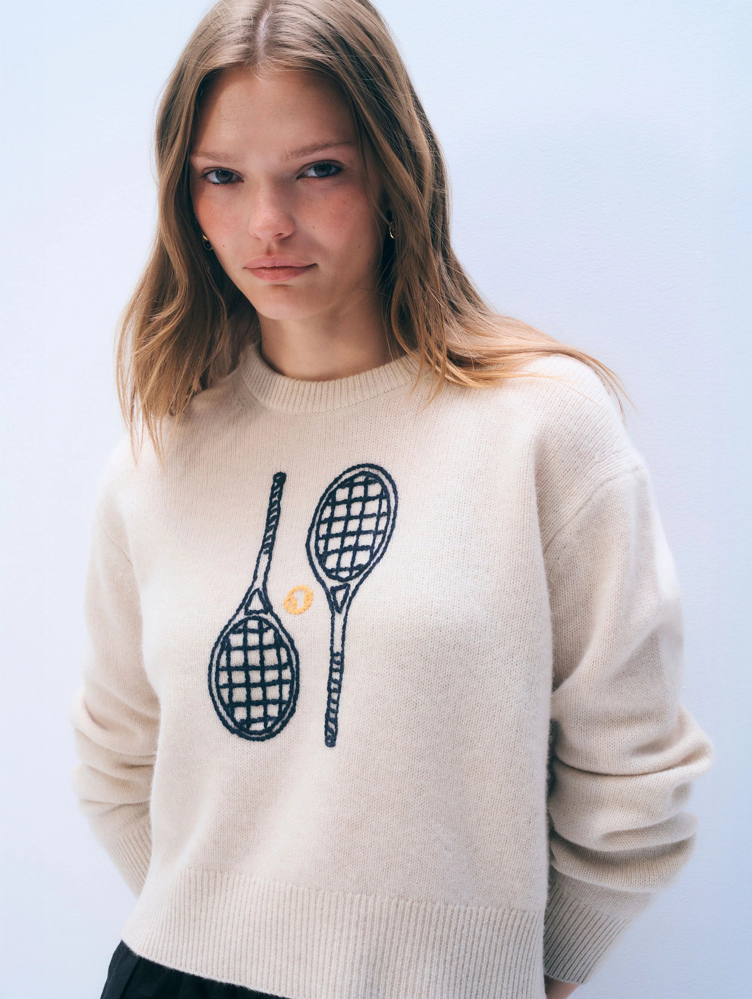 Woman wearing a cream sweater with tennis racket design on a light blue background