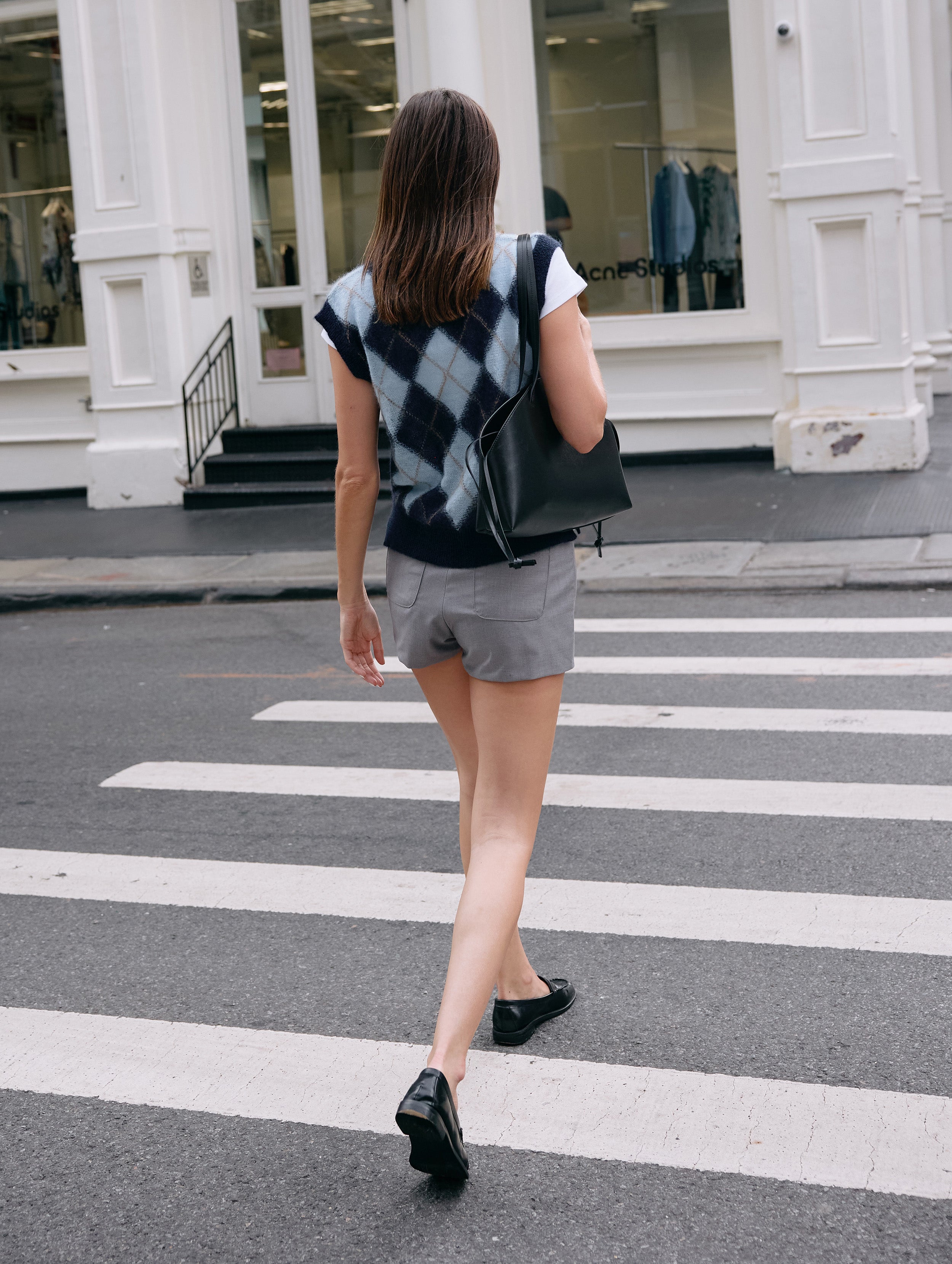Woman crossing a street wearing a sleeveless checkered top and gray shorts.