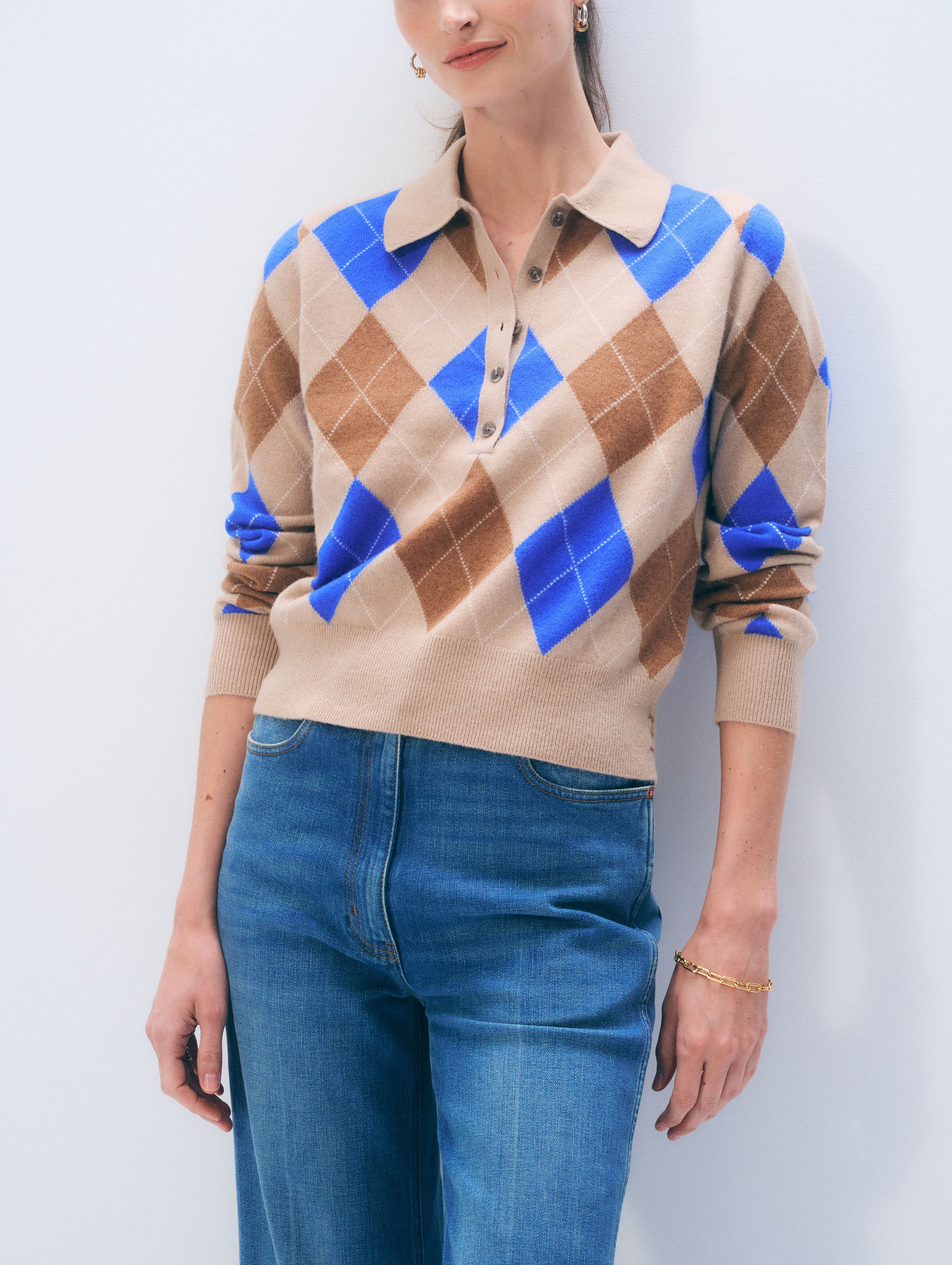 Person wearing a beige and blue argyle sweater with blue jeans against a white background