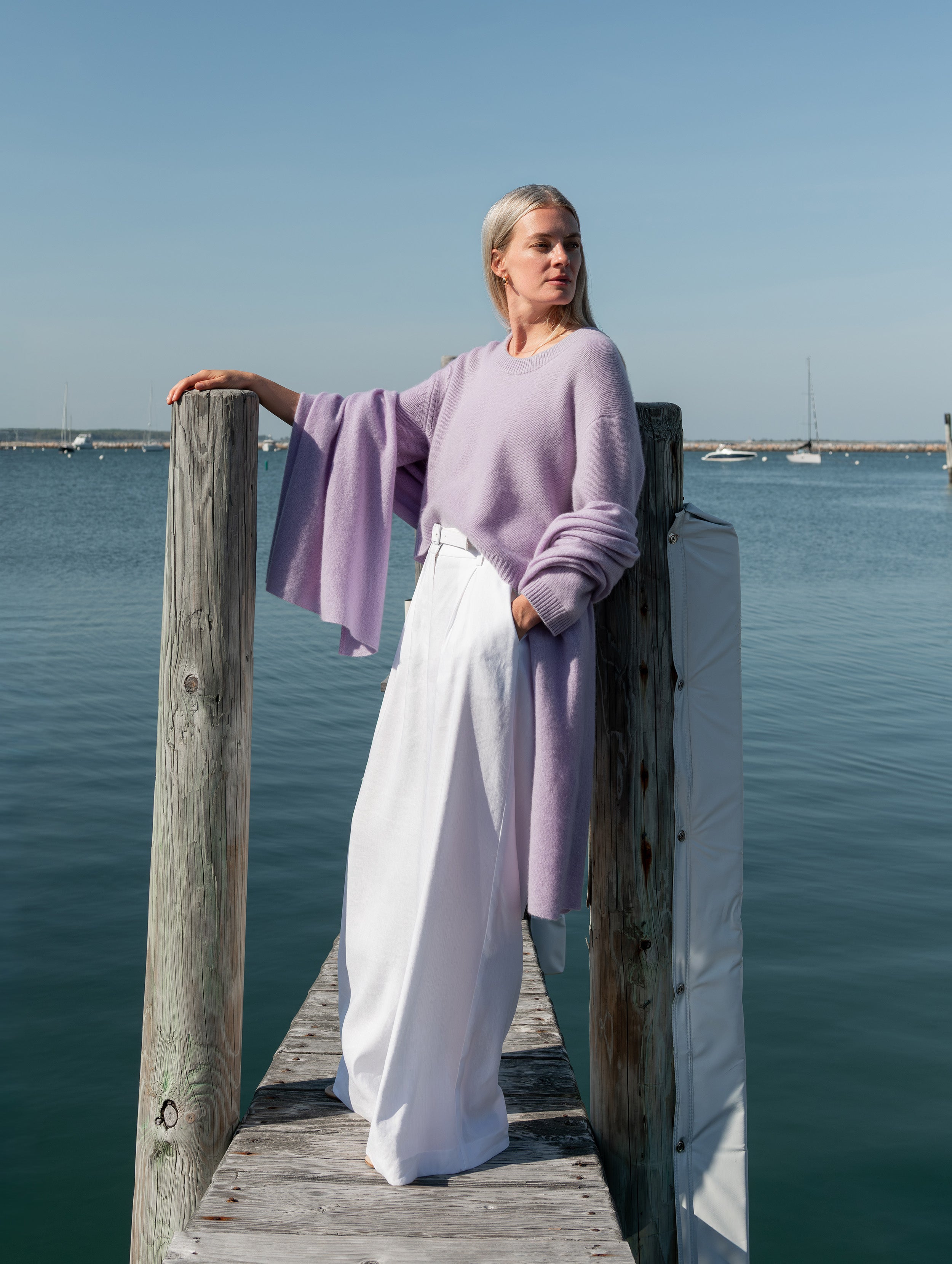 A woman stands on a wooden dock by the water wearing a lavender long-sleeve knit sweater and white wide-leg trousers.