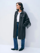 A woman stands against a plain light gray wall wearing a long charcoal gray fringed coat over a white top and blue jeans.
