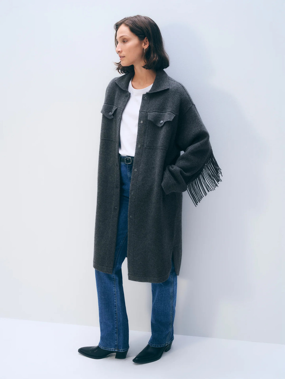 A woman stands against a plain light gray wall wearing a long charcoal gray fringed coat over a white top and blue jeans.