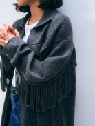 A person wearing a dark charcoal gray fringed cardigan over a white t-shirt and blue jeans, leaning against a light gray wall.