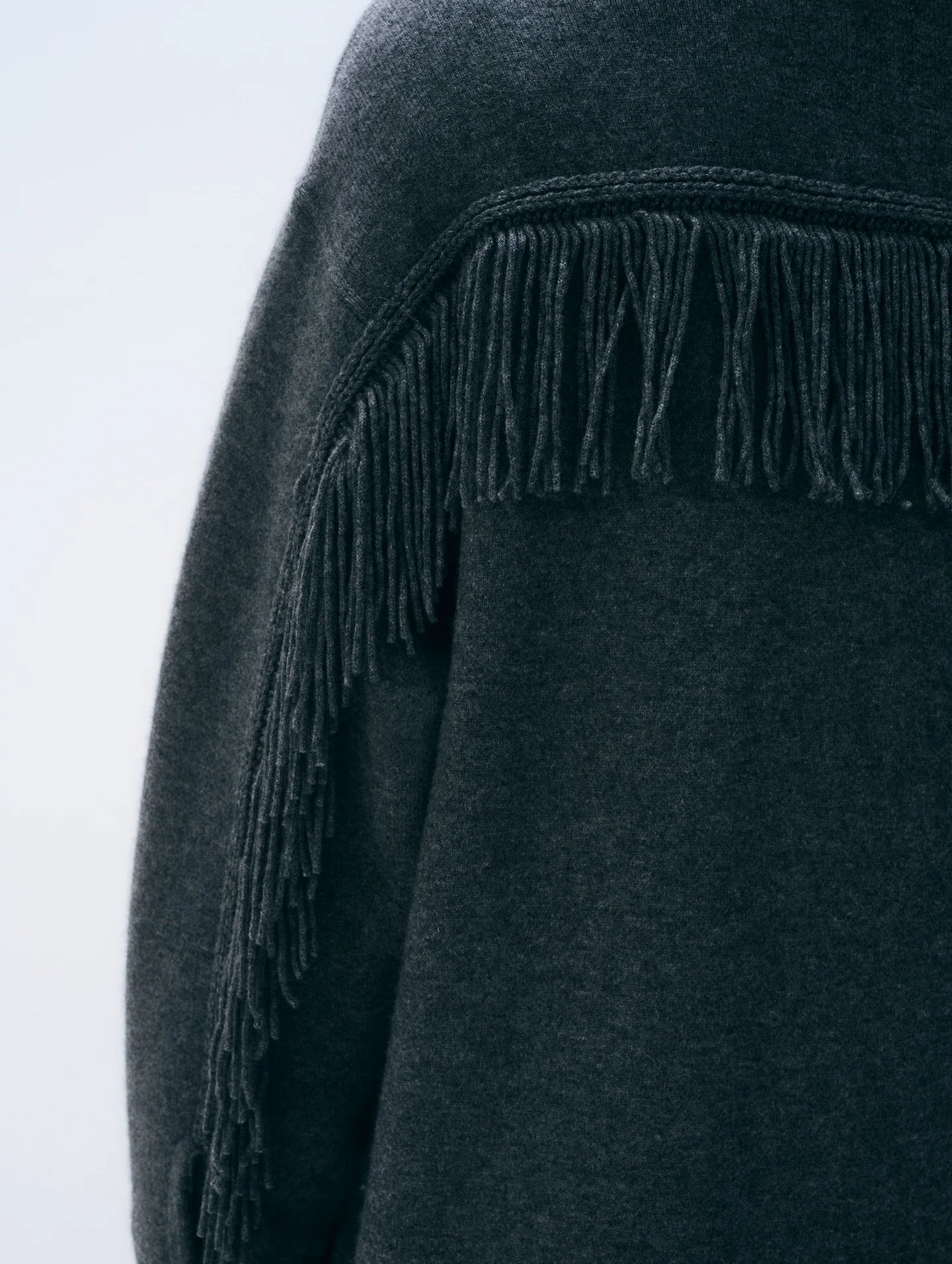 Close-up of a dark charcoal gray knit garment with long fringe trim along the shoulder, shown against a light gray background.
