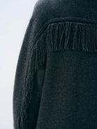 Close-up of a dark charcoal gray knit garment with long fringe trim along the shoulder, shown against a light gray background.