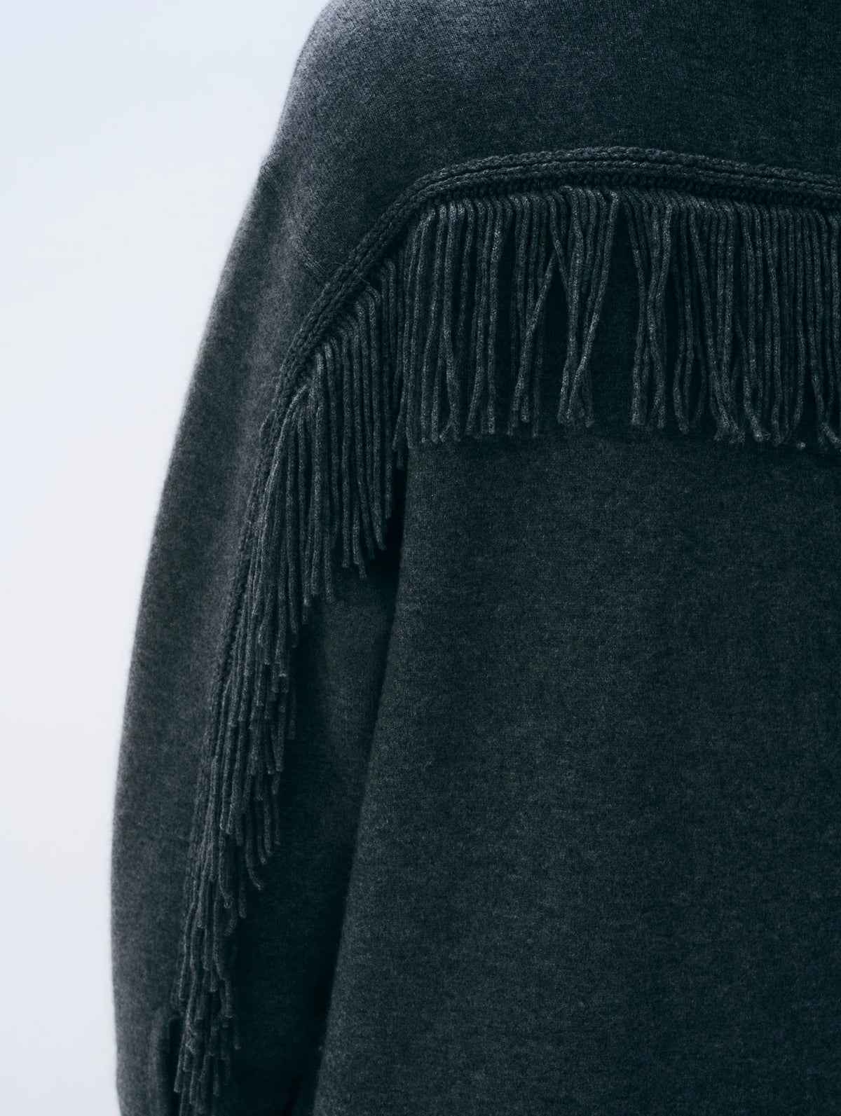 Close-up of a dark charcoal gray knit garment with long fringe trim along the shoulder, shown against a light gray background.
