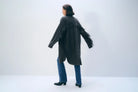 A woman is shown from the back wearing a dark charcoal gray fringed cardigan over blue jeans and black ankle boots, standing against a plain light gray studio background.